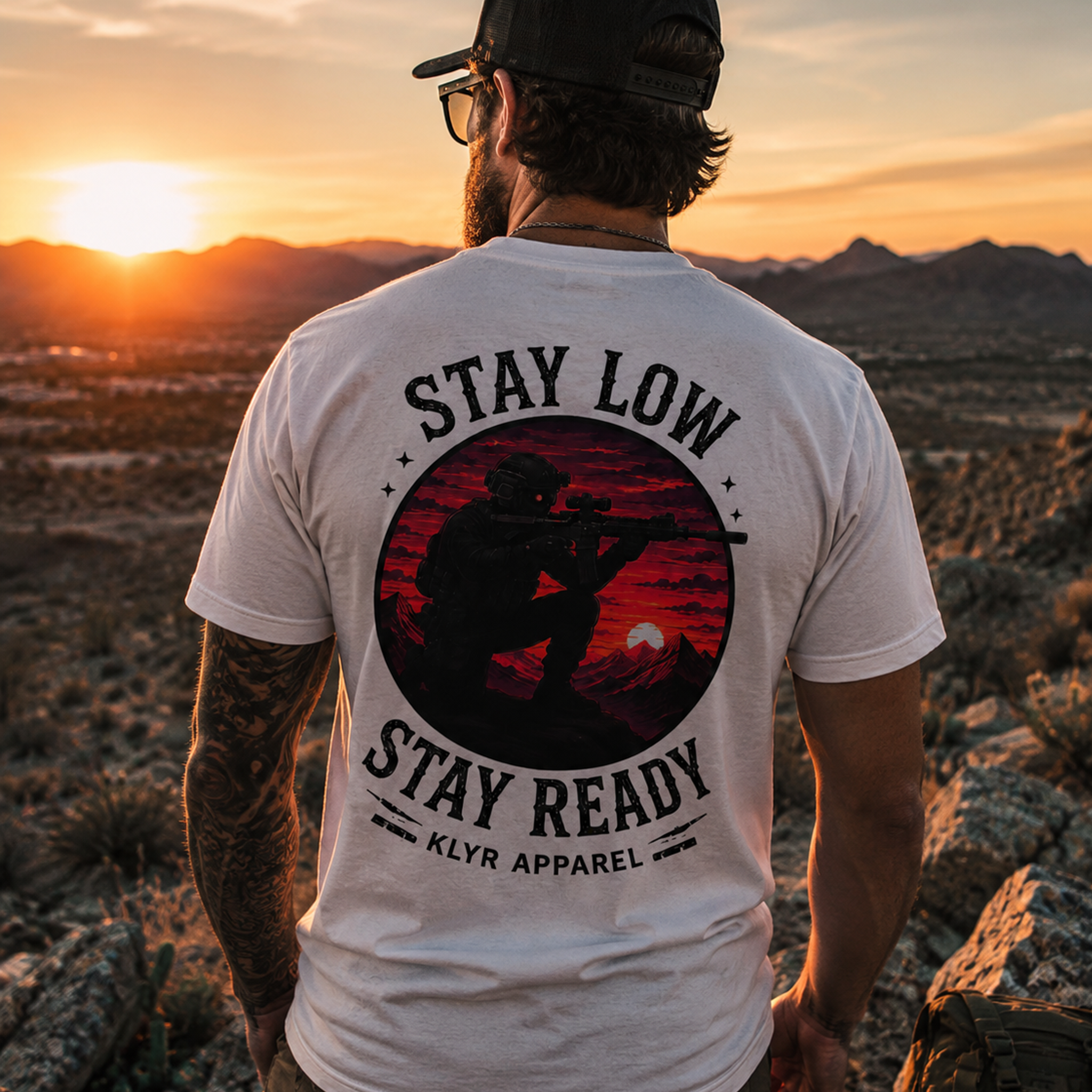 Person wearing the white KLYR Stay Low Stay Ready Tee showcasing the large tactical operator back graphic at sunset in the desert.