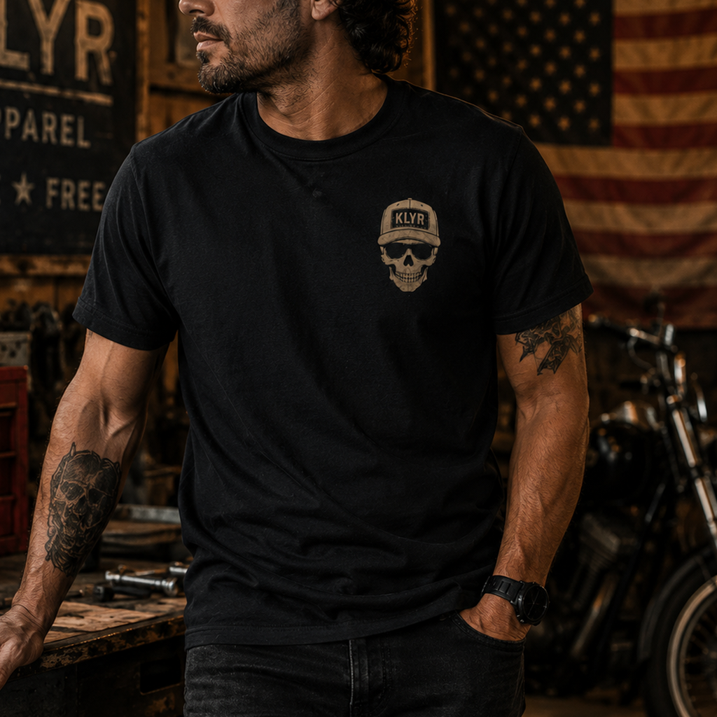 Front lifestyle view of the black KLYR Low Vis Tee featuring the skull and KLYR hat chest logo in a workshop setting.