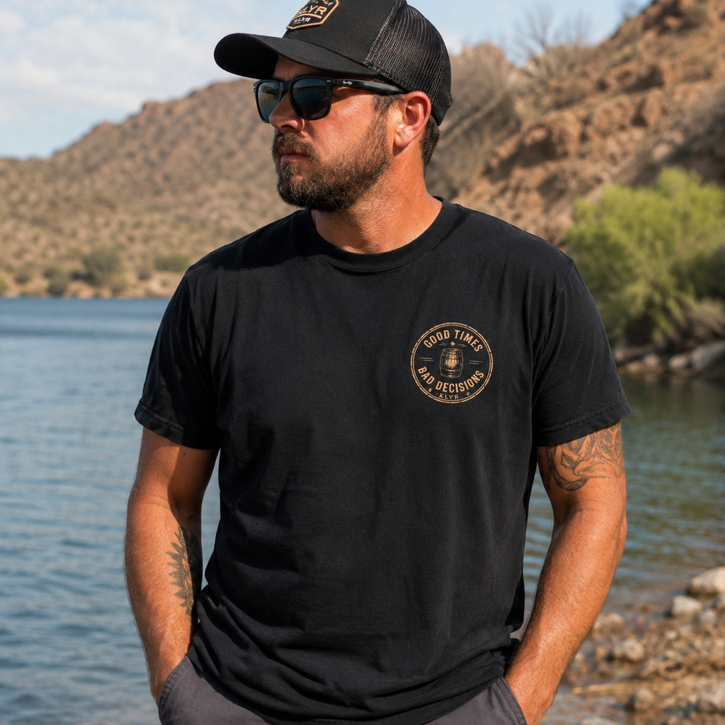 Front lifestyle view of the black KLYR Good Times Bad Decisions Tee featuring the vintage whiskey barrel chest logo at a desert lakeside.