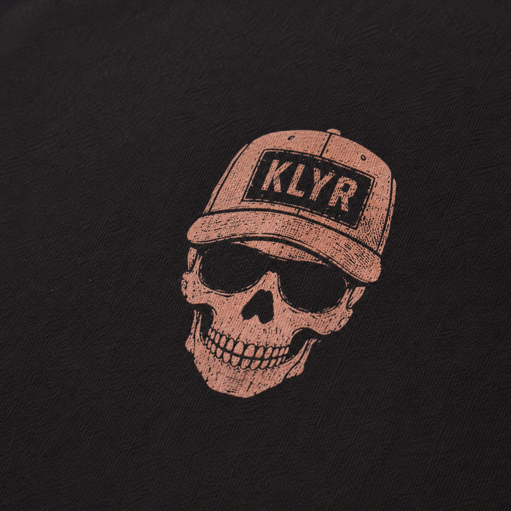 Detailed close-up of the Low Vis Tee chest logo featuring a skull wearing a KLYR trucker hat on the black KLYR t-shirt.