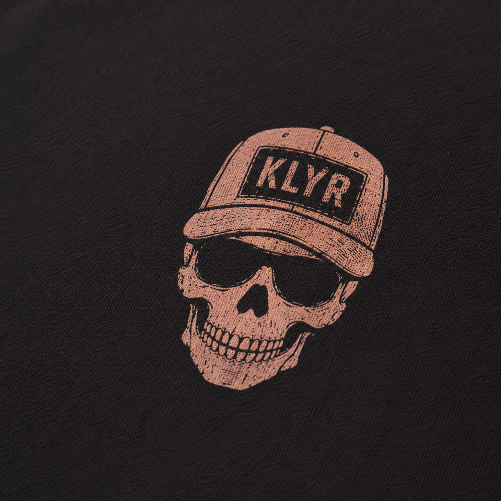 Detailed close-up of the Low Vis Tee chest logo featuring a skull wearing a KLYR trucker hat on the black KLYR t-shirt.