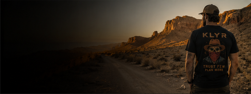 A man stands on a desert road at sunset wearing a black t-shirt with a large graphic on the back that reads 'KLYR' and 'TRUST FEAR PLAN MORE'.