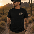 Person wearing the black KLYR Last Warning Tee with the cream KLYR chest badge visible against a desert landscape. 